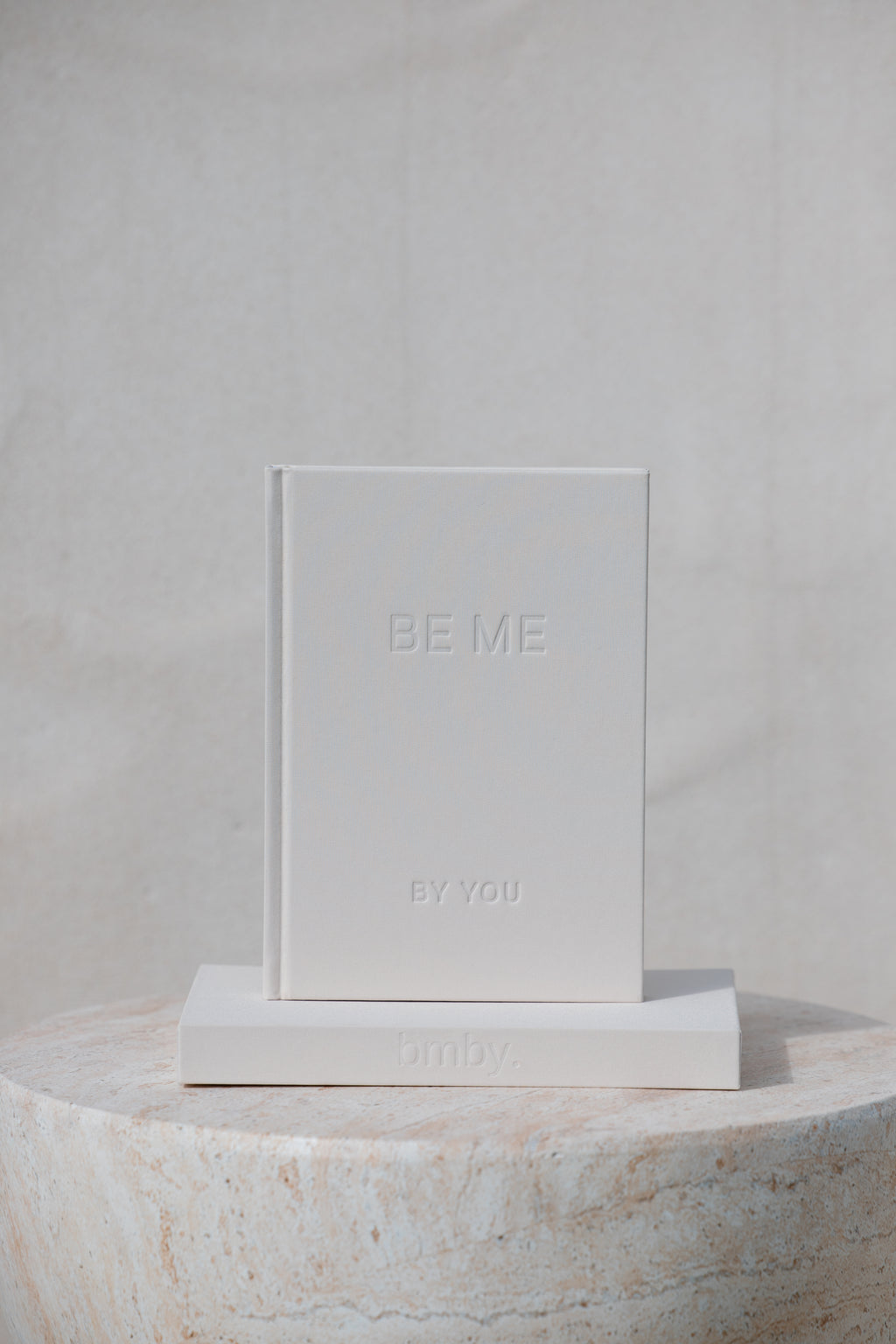 A minimalist white hardcover book or journal standing upright on a matching white rectangular base, with the embossed text 'BE ME' and 'BY YOU' visible on the cover. The product is photographed in a clean studio setup against a light neutral background, with part of a round stone pedestal visible at the bottom.