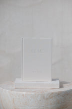 A minimalist white hardcover book or journal standing upright on a matching white rectangular base, with the embossed text 'BE ME' and 'BY YOU' visible on the cover. The product is photographed in a clean studio setup against a light neutral background, with part of a round stone pedestal visible at the bottom.