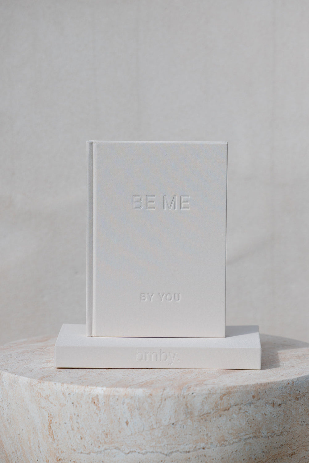 A minimalist white hardcover book titled 'BE ME BY YOU' with embossed lettering, displayed standing upright on a matching white block next to its front cover, set against a neutral textured beige background.