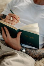 A close-up lifestyle shot of a person holding an open dark green journal with gold foil lettering that reads 'BE ME BY YOU'. The person is wearing a white t-shirt and beige pants, holding a green pen with a gold tip to write in the lined pages.