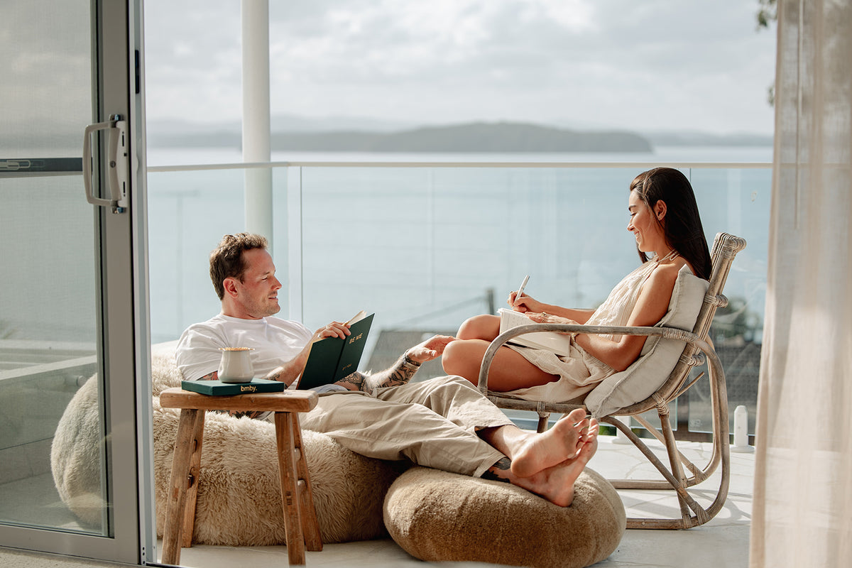 A man and a woman relax on lounge chairs on a sunlit balcony overlooking the ocean. The man wears a white shirt and beige pants while journalling in. BMBY BOOK, and the woman wears a light beige dress and holds a pen while journalling with bmby guided journal
