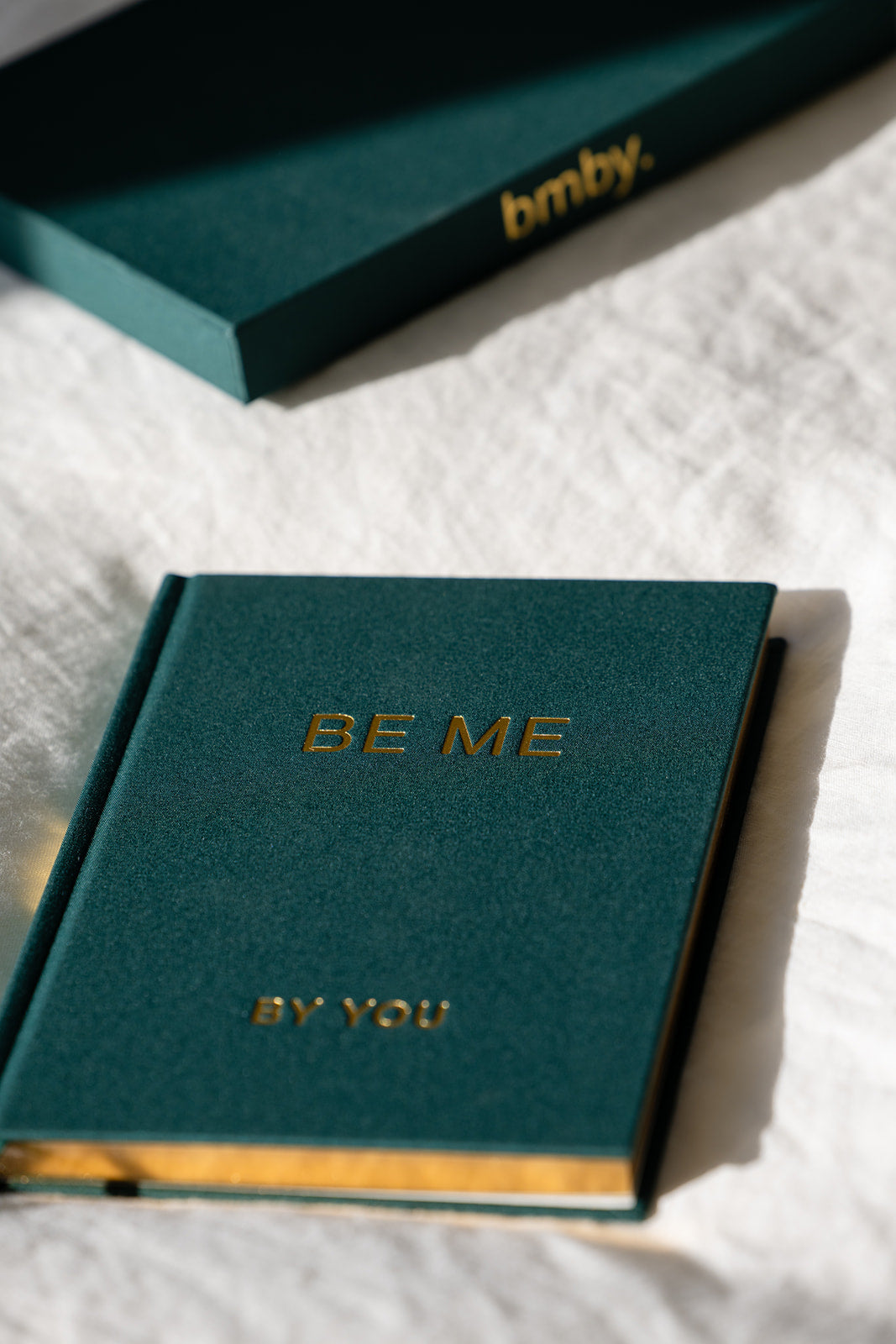 A dark teal hardcover journal with gold foil lettering reading 'BE ME' and 'BY YOU' on the cover, placed on a white textured fabric surface. A matching teal box with gold 'bmb.