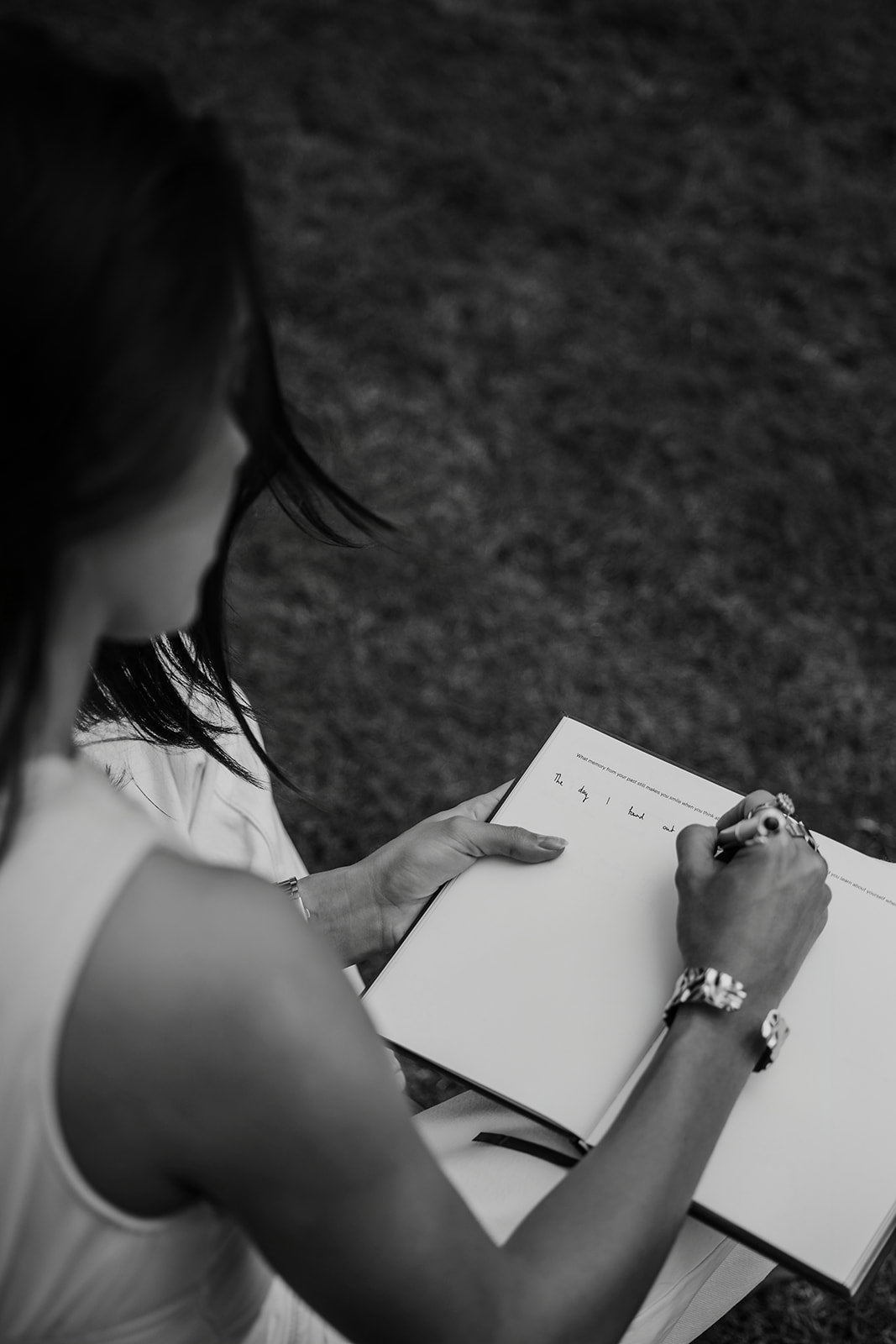 A black and white photograph of a woman writing in a large, open journal or book outdoors on grass.