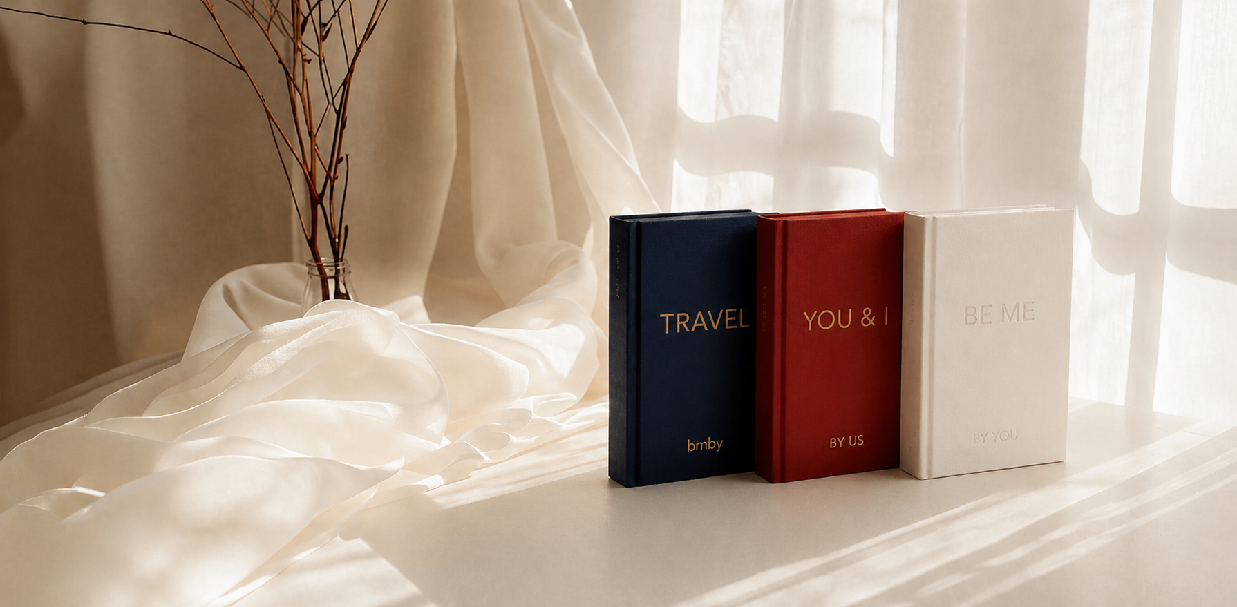 Three books titled 'TRAVEL', 'YOU & I', and 'WE' on a light surface with draped fabric and a plant in the background.